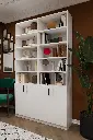 Beyaz ikili kitaplık, açık raflarda kitaplar ve objeler; alt bölümde kapaklı dolaplar, yanında yeşil bitki.